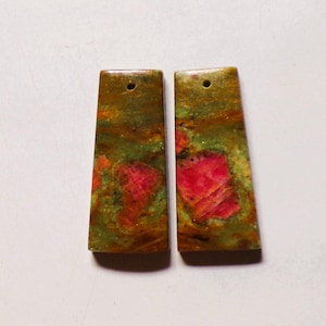 Peut inclure: Deux pendentifs en pierre rectangulaires présentant une surface marbrée avec des tourbillons de vert olive, de brun terreux et d'inclusions rose magenta vif. Chaque pièce plate possède un petit trou circulaire près du bord supérieur.