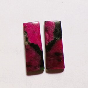 Könnte beinhalten: Zwei rechteckige Cabochons mit einem lebendigen magentafarbenen und tiefschwarzen Marmormuster. Diese polierten Steine haben flache Oberflächen und gerade Kanten und zeigen eine kontrastreiche Farbkombination mit organischen, dunklen Adern.