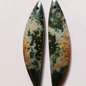 May include: Two polished, teardrop-shaped gemstones with a hole drilled at the top for jewelry making. The stones feature a mottled pattern of dark green, tan, and light blue colors. The bottom edges are a deep, dark green.