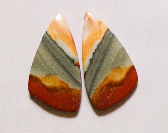 May include: Two teardrop-shaped polished gemstones featuring horizontal bands of color. The stones display layers of cream, soft gray, golden yellow, and deep terracotta red, with a smooth, glossy finish and distinct natural mineral patterns.