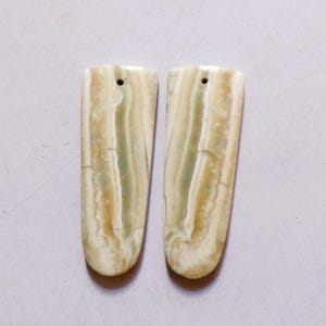 Può includere: Due pendenti allungati in pietra lucida con un foro nella parte superiore per l'infilatura. I pendenti presentano un motivo a bande color crema, verde e beige. Il fondo arrotondato e la forma rettangolare conferiscono loro un aspetto unico.