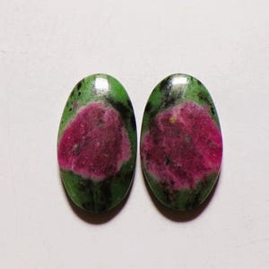 May include: Two oval-shaped cabochons featuring a vibrant magenta-pink centre surrounded by a deep green matrix with black inclusions. These polished gemstones have a smooth, reflective surface and a distinct colour contrast between the pink and green.