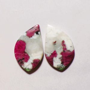 Könnte beinhalten: Zwei tropfenförmige Cabochons mit polierter Oberfläche und einem gesprenkelten Muster aus leuchtendem Magenta, opakem Weiß sowie kleinen Einschlüssen von dunkelgrünen und schwarzen Mineralien im Steinmaterial.