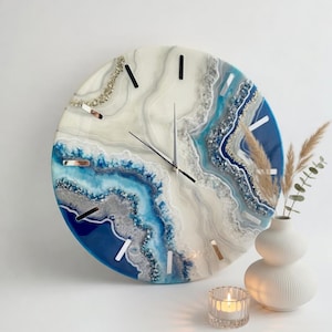 Può includere: Un orologio da parete rotondo con un design ispirato ai geodi, caratterizzato da strati vorticosi di resina bianca, crema e varie tonalità di blu. Il quadrante presenta indici argentati e sottili lancette color argento con dettagli cristallini.