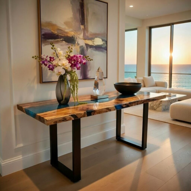 May include: A wooden console table with a blue resin inlay and black metal legs. The table is decorated with a vase of flowers, a crystal sculpture, and a black bowl. The background features a painting and a view of the ocean.
