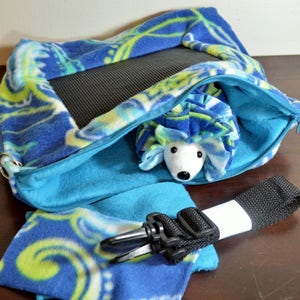 May include: A blue and green patterned fabric hedgehog sleeping bag with a zipper closure and a black strap. The sleeping bag is open and a stuffed hedgehog is inside.