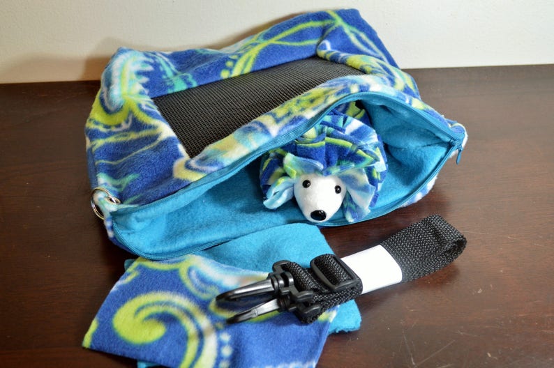May include: A blue and green patterned fabric hedgehog sleeping bag with a zipper closure and a black strap. The sleeping bag is open and a stuffed hedgehog is inside.