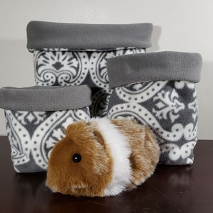 May include: Three soft fabric storage bins in varying sizes featuring a grey and white damask pattern with solid grey folded rims. A plush guinea pig toy with brown and white fur is positioned in front of the bins on a dark surface.