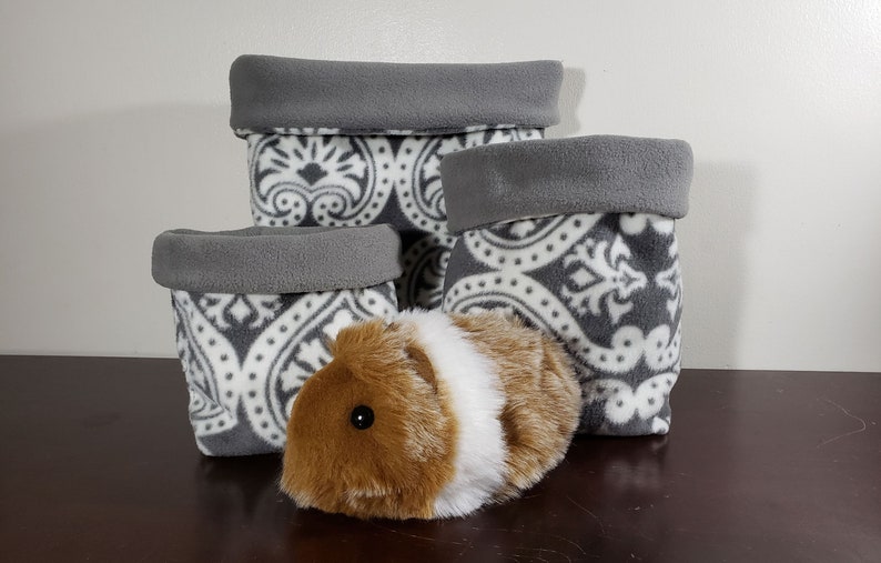 May include: Three soft fabric storage bins in varying sizes featuring a grey and white damask pattern with solid grey folded rims. A plush guinea pig toy with brown and white fur is positioned in front of the bins on a dark surface.