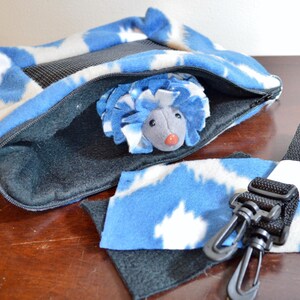 May include: A blue and white fleece hedgehog carrier with a zippered opening and a black strap with clips. The carrier has a mesh panel for ventilation. A gray plush hedgehog toy is inside the carrier.