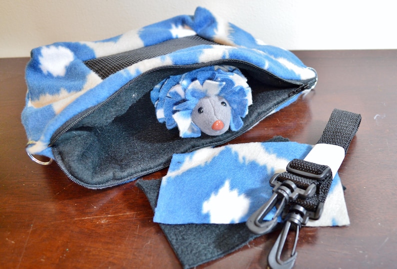 May include: A blue and white fleece hedgehog carrier with a zippered opening and a black strap with clips. The carrier has a mesh panel for ventilation. A gray plush hedgehog toy is inside the carrier.