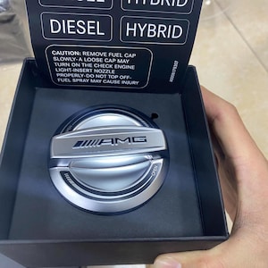 May include: A silver and black fuel cap with the AMG logo, displayed in a black box. The cap has the word "OPEN" on it. The text "DIESEL" and "HYBRID" are visible above the cap. The box also contains a warning about removing the fuel cap.