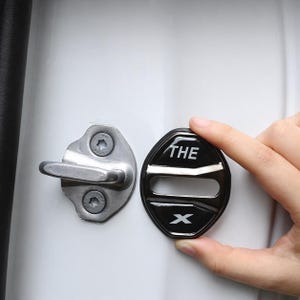 Puede incluir: Una cubierta negra para la cerradura de la puerta del coche con la palabra "THE" y una "X" en blanco. La cubierta se sostiene cerca de un pestillo plateado de la puerta del coche en una puerta blanca.