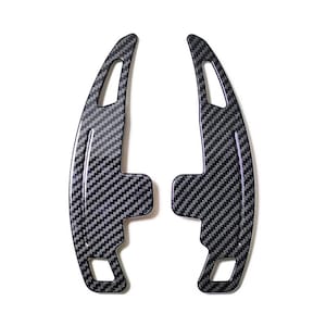 Puede incluir: Dos levas de cambio de fibra de carbono con un patrón tejido en negro y gris. Las levas tienen un diseño curvo con forma de paleta y una base rectangular. El fondo es blanco.