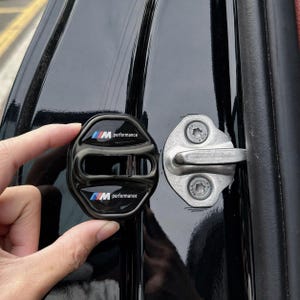 Puede incluir: Cubierta negra para cerradura de puerta de coche con el texto "M performance" en blanco, azul y rojo. La cubierta se sostiene junto a un pestillo plateado de puerta de coche. La puerta del coche es negra.