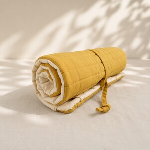 May include: A mustard yellow quilted blanket rolled into a cylinder shape. The blanket features a textured, grid-like stitched pattern on the exterior and a smooth, cream-colored interior lining. A matching mustard yellow fabric tie secures the roll.