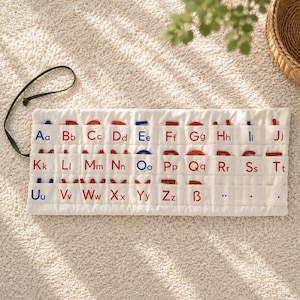 May include: A white fabric alphabet organizer featuring three horizontal rows of pockets. Each pocket is labeled with an uppercase and lowercase letter in red or blue embroidery. Red and blue wooden letters are tucked into the pockets.