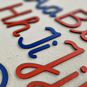 Op de afbeelding: Een verzameling individuele, driedimensionale letters in rood en blauw, gerangschikt op een gestructureerde, gebroken witte stoffen ondergrond. De letters hebben een gladde, matte afwerking en zijn uitgesneden in een cursief lettertype.