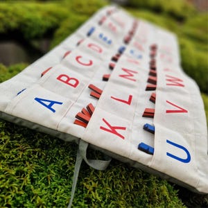 May include: A canvas alphabet organizer with red and blue letters. The organizer is filled with wooden letter tiles. The letters are printed on individual pockets. The background is a bed of green moss.