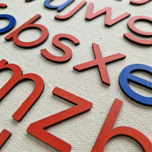 May include: Close-up of wooden alphabet letters in red and blue. The letters are scattered on a neutral-colored fabric surface. The letters are in a variety of fonts and sizes, suitable for educational or decorative purposes.