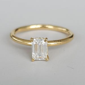 May include: A gold ring with a rectangular, clear gemstone. The ring has a thin band and a four-prong setting. The gemstone is the focal point, reflecting light. The ring is set against a neutral background. The text "VEXA JEWEL" is visible.