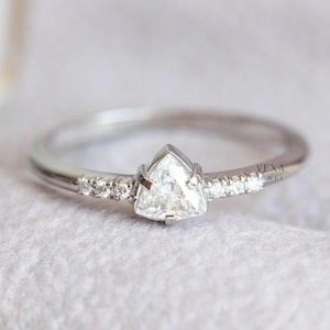 May include: A silver ring featuring a triangular, clear gemstone as the centerpiece. Small, round gemstones are set along the band. The ring is set against a soft, white background. The text "VEXA JEWEL" is visible.