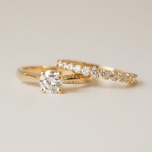 May include: A pair of gold rings. One ring has a large, round, clear gemstone. The second ring is a band with multiple smaller, clear gemstones. The rings are on a white surface. The text "VEXA JEWEL" is visible.