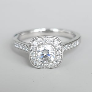 May include: A silver ring featuring a large, clear, round central stone encircled by smaller, clear stones in a square halo. The band is also set with small, clear stones. The ring is set against a light grey background.