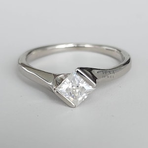 May include: A silver ring with a square, clear gemstone. The ring has a modern design with a split shank and a bezel setting. The text "Vexa Jewel" is visible on the side of the ring.