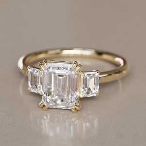 May include: A gold engagement ring featuring a large emerald-cut central diamond flanked by two smaller stones. The ring has a polished gold band. The diamonds are clear and reflect light.