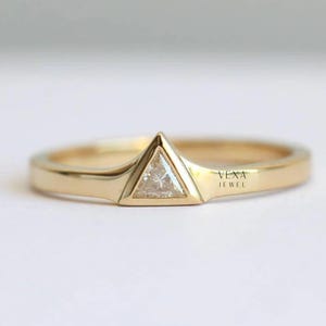 May include: A gold ring with a triangular diamond center stone. The ring has a smooth, polished band and a unique setting that highlights the geometric shape of the diamond. The text "VEXA JEWEL" is visible on the ring.