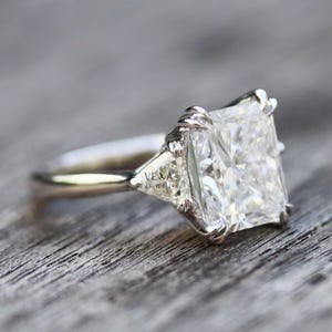 May include: A radiant-cut diamond engagement ring with a platinum band. The ring features a large, square-cut center stone and two smaller, triangular side stones. The ring is set on a wooden surface. The text "Vexa Jewel" is visible.