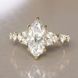 May include: A gold engagement ring with a marquise-cut diamond centre stone. The band is adorned with smaller, round diamonds. The ring is set against a neutral background. The ring is a piece of jewellery.