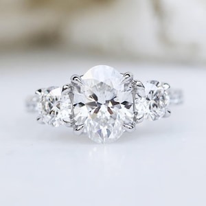 May include: A three-stone engagement ring with a large oval-cut center diamond and two smaller side stones. The ring is set in a silver band and features the text "Vexa Jewel". The diamonds sparkle brightly against a white background.