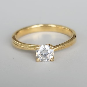 May include: A gold solitaire engagement ring featuring a round, clear diamond. The ring has a classic design with a polished band. The ring is set against a neutral background. The ring is stamped with the text "Vexa Jewel".