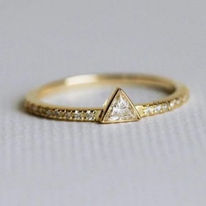 May include: A gold ring with a triangular, clear gemstone at the center. The band is also gold and is encrusted with small, clear gemstones. The ring is set against a soft, neutral background. The text "VEXA JEWEL" is visible.