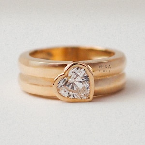 May include: A gold ring with a heart-shaped diamond. The ring has a thick band with a polished finish. The diamond is set in a bezel setting, and the ring is displayed on a white background. The text "VEXA JEWEL" is visible on the ring.