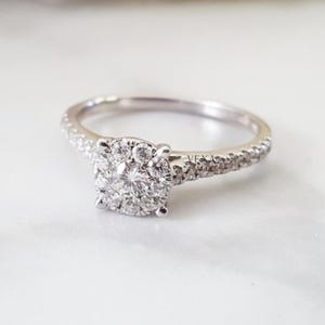 May include: A diamond engagement ring with a cluster of small diamonds at the center and smaller diamonds set along the band. The ring is set in a white metal, likely platinum or white gold, and sits on a white surface.