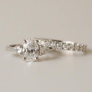 Peut inclure: Deux bagues en diamant. La bague de fiançailles présente un diamant de taille ovale flanqué de deux diamants en forme de poire. L'alliance est une bague délicate avec plusieurs petits diamants. Les deux bagues sont en métal argenté.