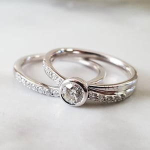 May include: Two silver rings. One ring has a large, round, clear gemstone in a bezel setting. The second ring is a thin band with small, clear gemstones. The rings are on a white surface.