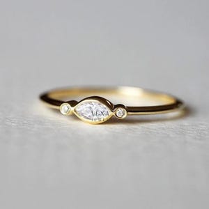 May include: A delicate gold ring featuring a marquise-cut diamond center stone, flanked by two smaller round diamonds. The ring has a thin, polished band and the text "VEXA JEWEL" is visible.