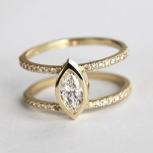 May include: A gold-colored ring with a marquise-cut diamond center stone. The ring features a split shank design, with a band of small diamonds on each band. The ring is set against a white background.