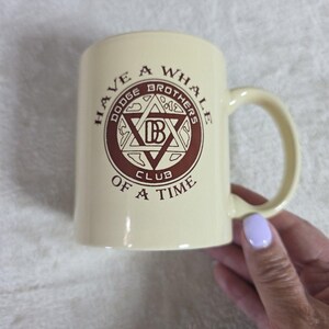 May include: Cream-colored ceramic mug with a handle. The mug features a circular emblem with the text "Dodge Brothers Club" and the phrase "Have a Whale of a Time" in brown lettering. The mug is held by a hand with a light purple nail polish.