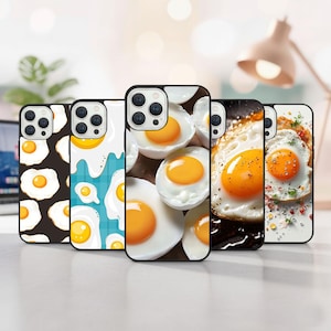 以下が含まれることがあります： 目玉焼きのデザインが施された携帯電話ケースのコレクション。ケースには、暗い背景の卵、青と白のパターン、調理された卵のクローズアップなど、さまざまなパターンが表示されています。ケースは黒い縁取りで、明るい表面に配置されています。