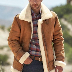May include: A brown shearling jacket with a cream-colored shearling lining and trim. The jacket features a zip-up front, a wide collar, and a plaid shirt underneath. The person is wearing blue jeans and a brown belt.