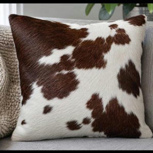 Könnte beinhalten: Quadratisches Zierkissen mit Kuhfellmuster. Das Kissen hat ein braun-weißes Fleckendesign, das an ein Kuhfell erinnert. Es liegt auf einem grauen Sofa und verleiht der Einrichtung einen rustikalen Touch.