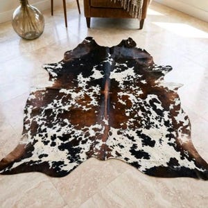 Puede incluir: Una alfombra de piel de vaca con una forma orgánica irregular y un patrón moteado de manchas de color marrón oscuro, negro y blanco. La piel muestra variaciones de color naturales en toda su superficie, creando un contraste marcado.