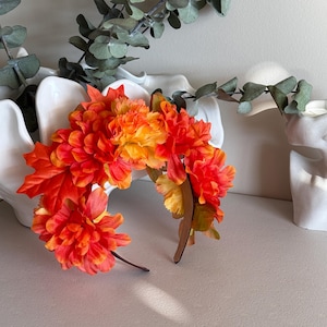 May include: A vibrant floral headband featuring orange and yellow artificial flowers and leaves. The headband is made of a brown material and is adorned with a variety of colorful blooms, perfect for adding a touch of nature-inspired flair to any outfit.