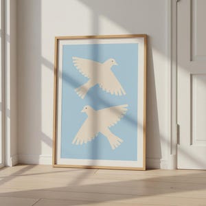 Peut inclure: Une affiche encadrée représentant deux colombes crème en vol sur un fond bleu clair. L'œuvre est encadrée dans un cadre en bois naturel, créant une esthétique minimaliste et sereine. L'affiche est appuyée contre un mur blanc.