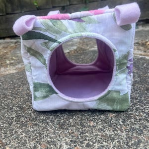 Puede incluir: Un escondite para mascotas en forma de cubo suave con un exterior de tela blanca con un estampado floral verde y morado. El interior está forrado con tela de color morado claro sólido y las esquinas superiores tienen pequeñas presillas.